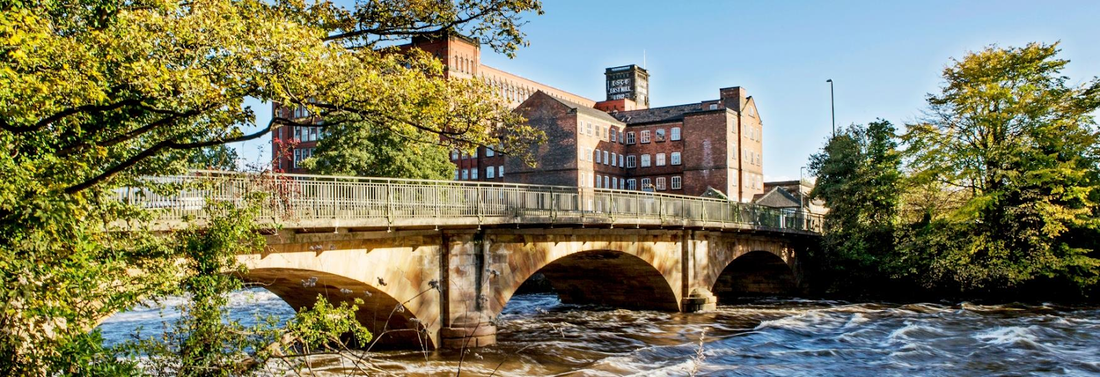 A bridge crossing the fast-moving Derwent river, with the large brick building in the background of Derwent Valley Mills on a sunny day.