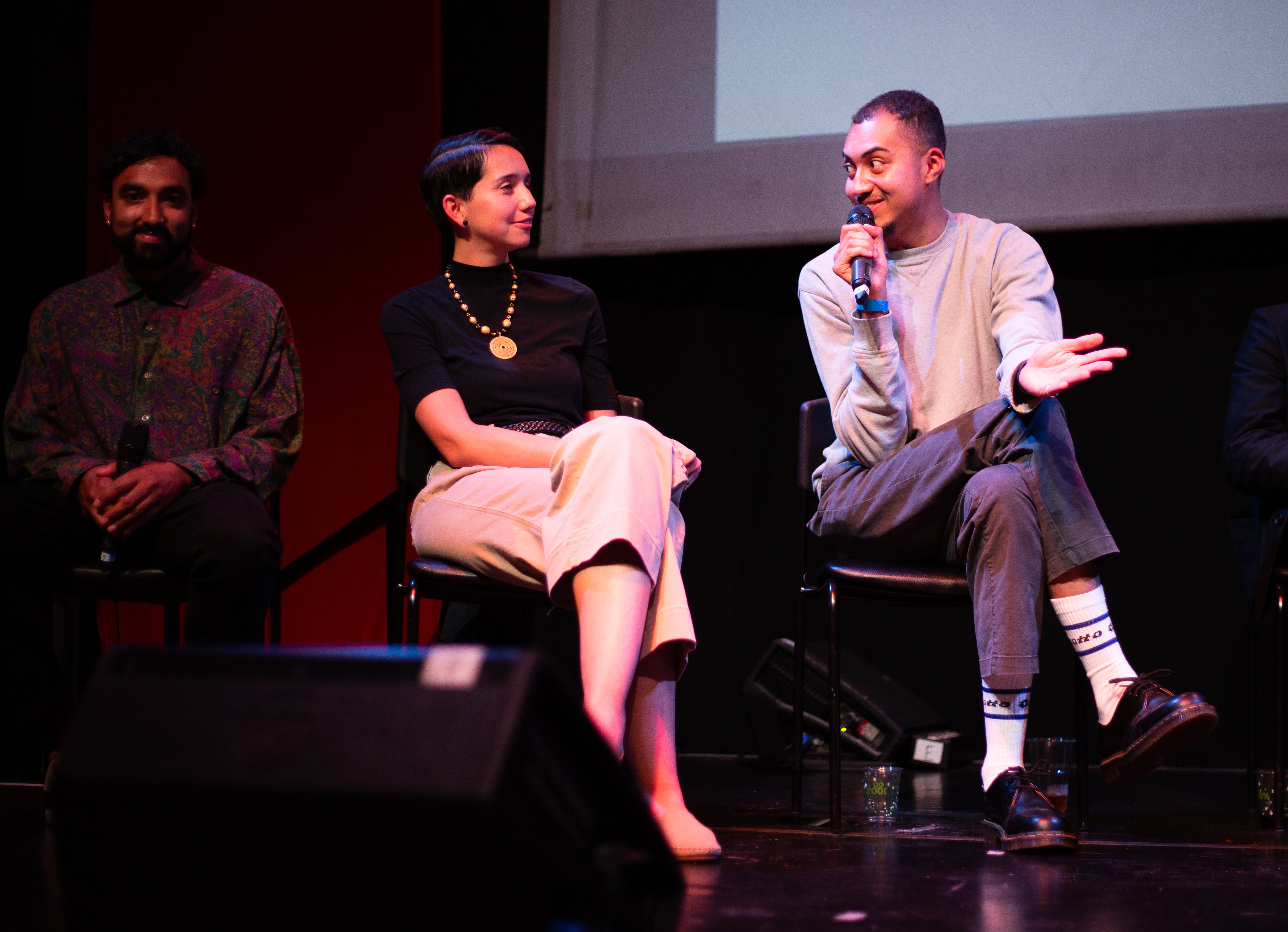 three people sit on a dark stage lit with pink, with a projected screen behind them. Louis sits on the right holding the mic and talking expressively
