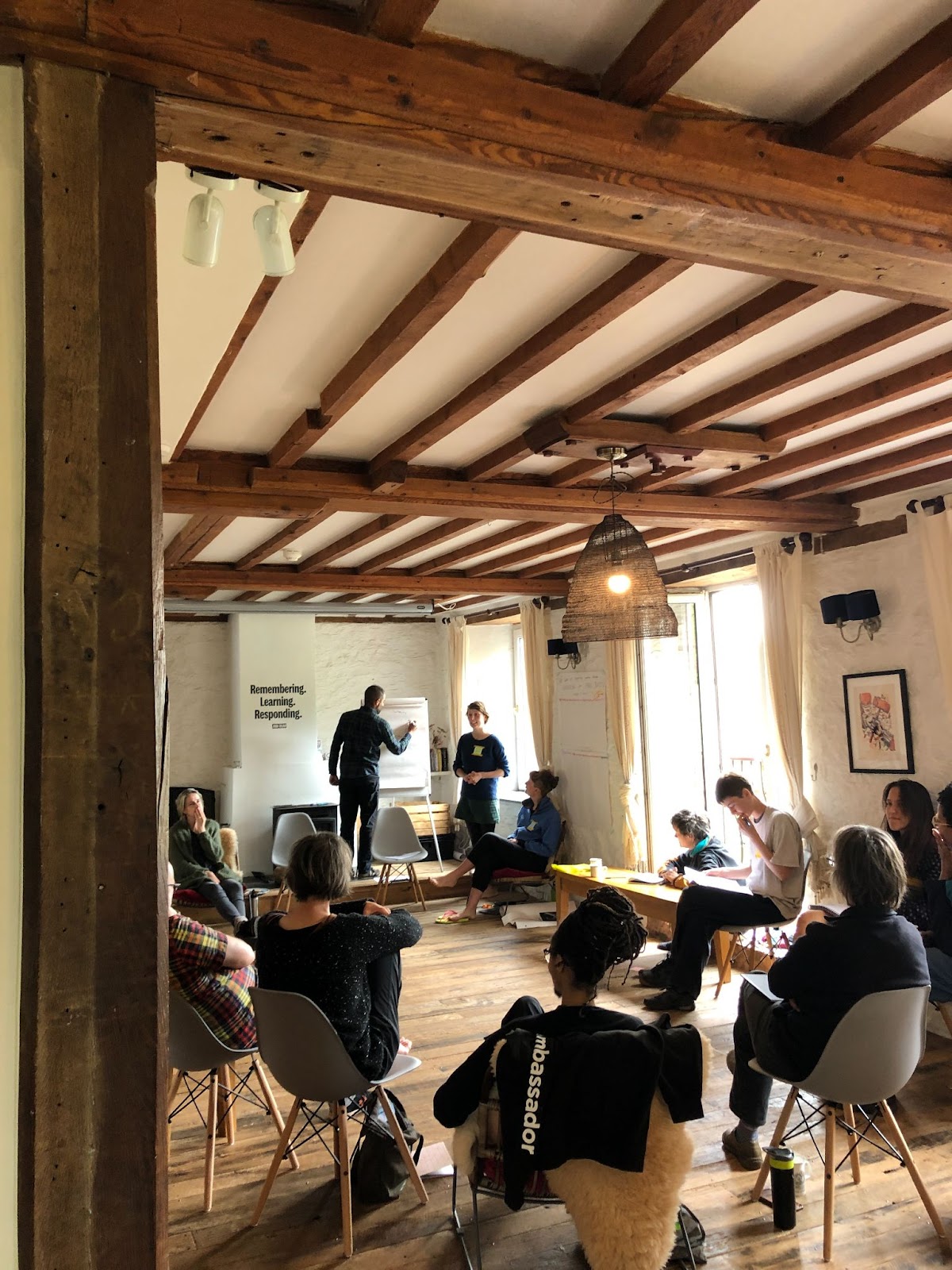 Inside the mill house at Selgars Mill, a group of people sit in a circle of chairs, with two people standing and one of them writing on a flipchart.