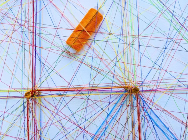 Network of multicoloured strings threaded across a white background. One spool of orange string is visible under the strings.