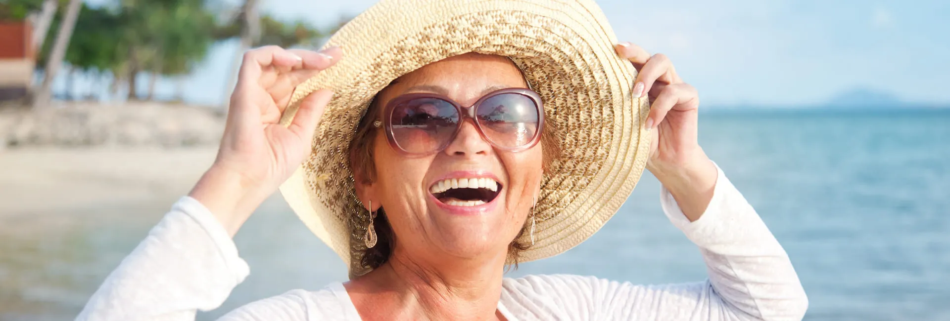 Female senior traveller, smiling, basking in the sun with sunglasses and a straw hat, enjoying a moment by the ocean at the beach.