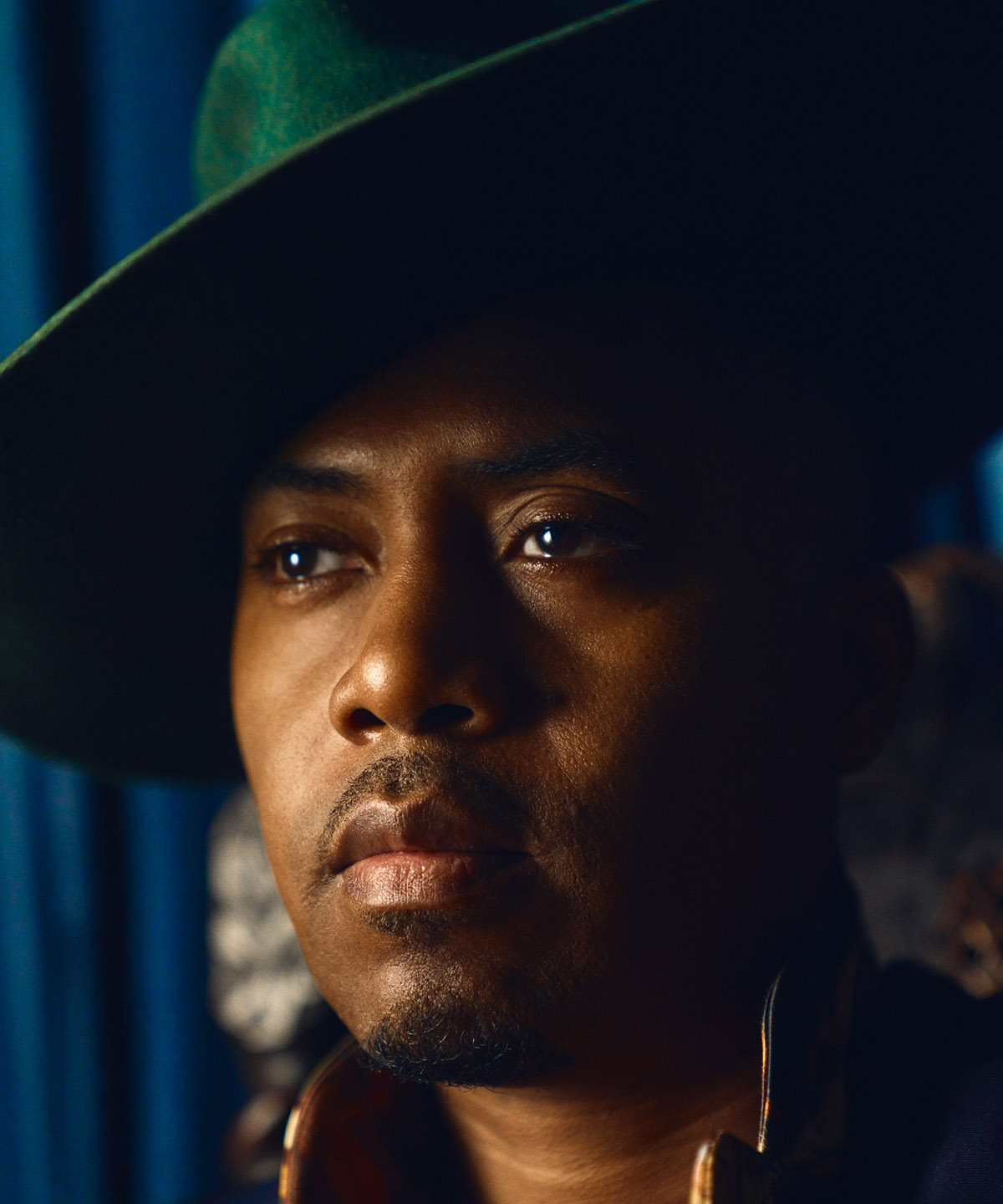 up close photo of nas wearing green hat with a blue background lightly blurred