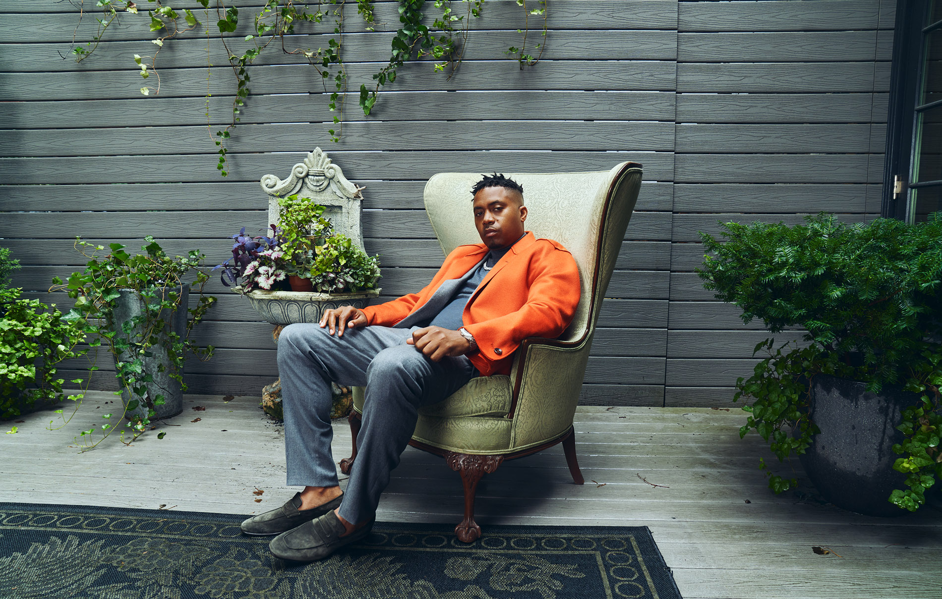 nas wearing an orange jacket sitting in a chair looking relaxed with three green plant beside him and wooden fence in the background