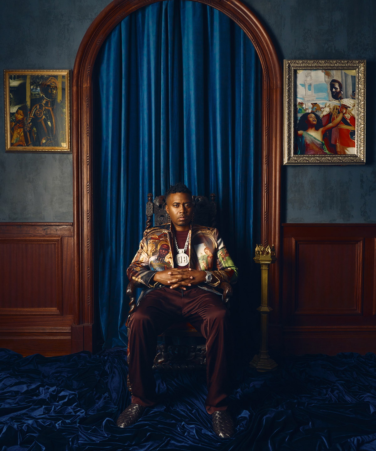 nas sitting in a big chair with two piece of art work beside him with a blue curtain background