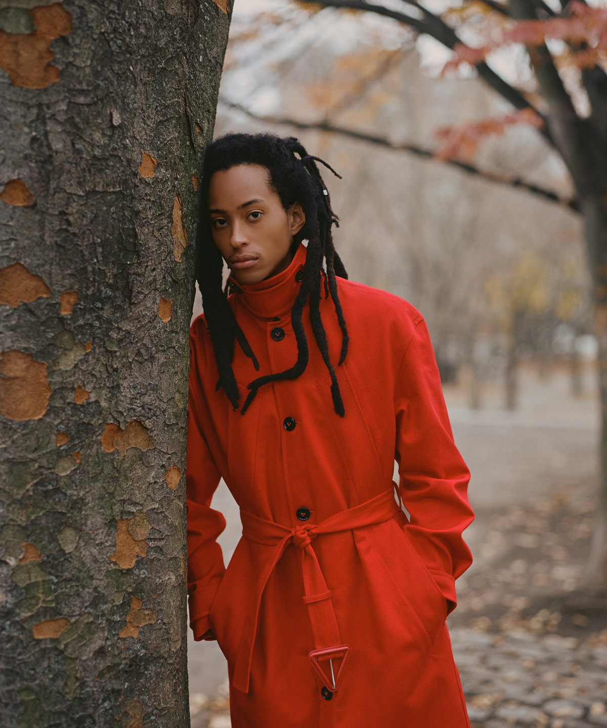 long haired model wearing red jacket leaning on a tree