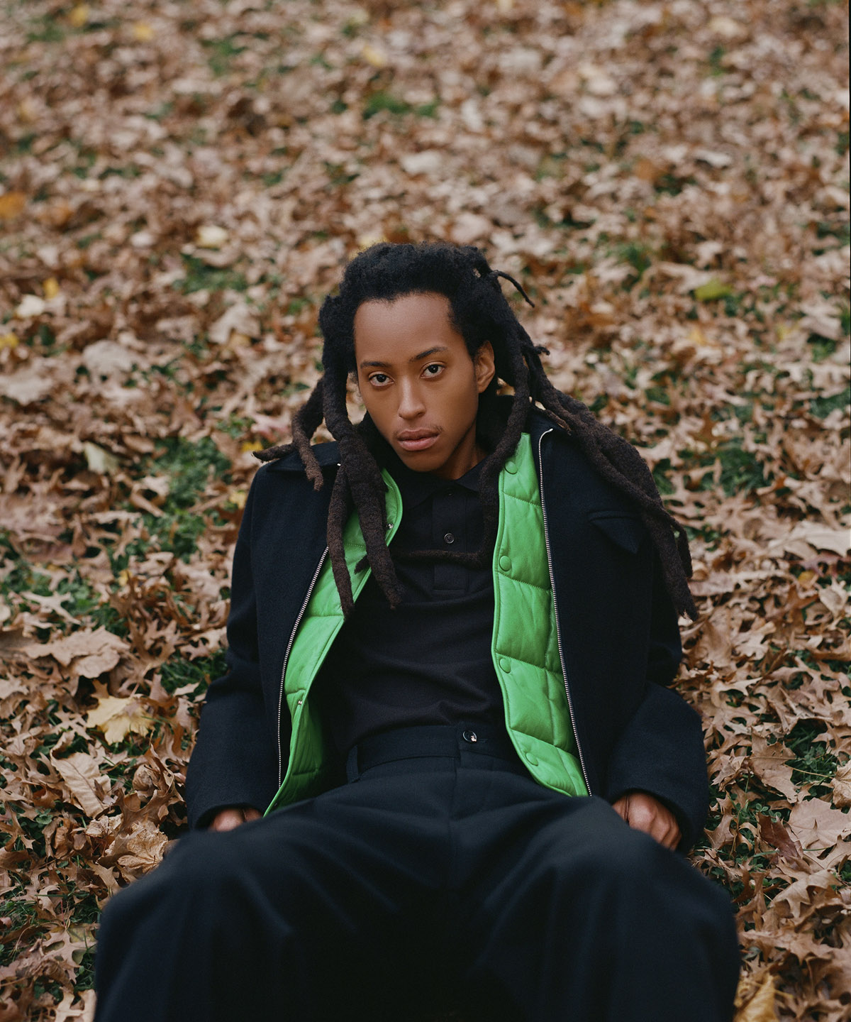 model sitting on the ground wearing black pants black shirt and a green vest with many leaves on the ground