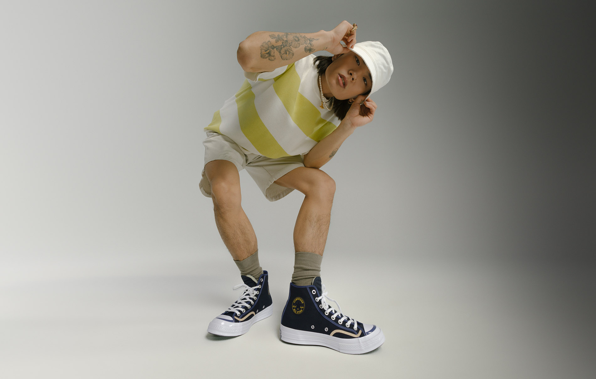 male model wearing bucket hat with bent down and hand holding his hat while displaying his converse shoe