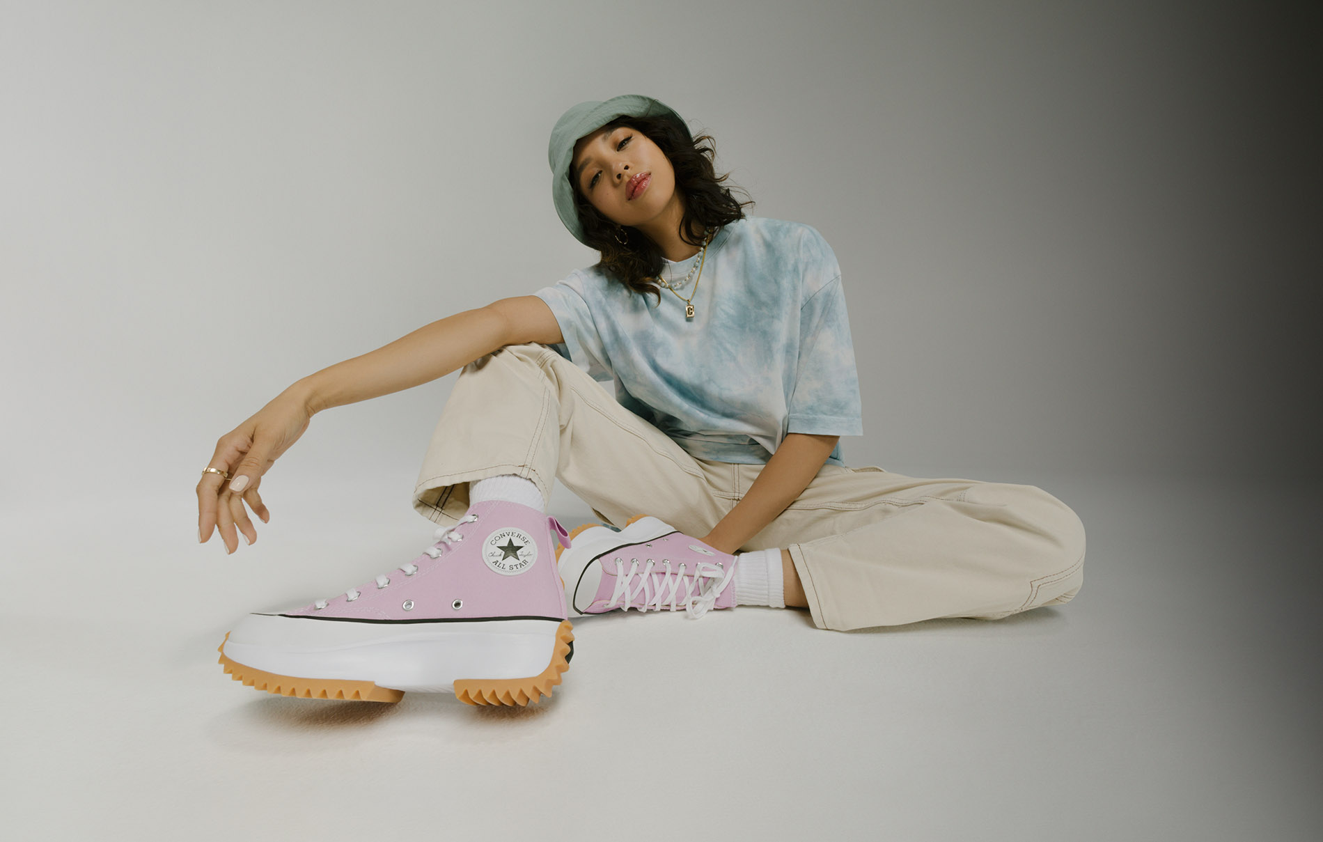 women model sitting down wearing pink converses with a tie dye shirt and bucket hat