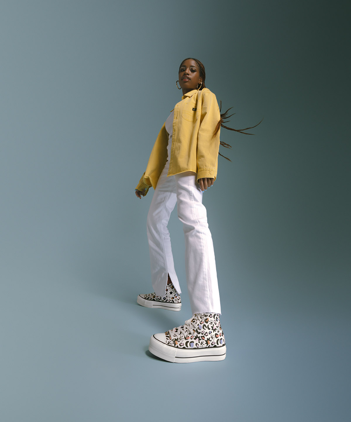 Women model wearing yellow jacket and white pants with cheetah print converse shoes in a blue background