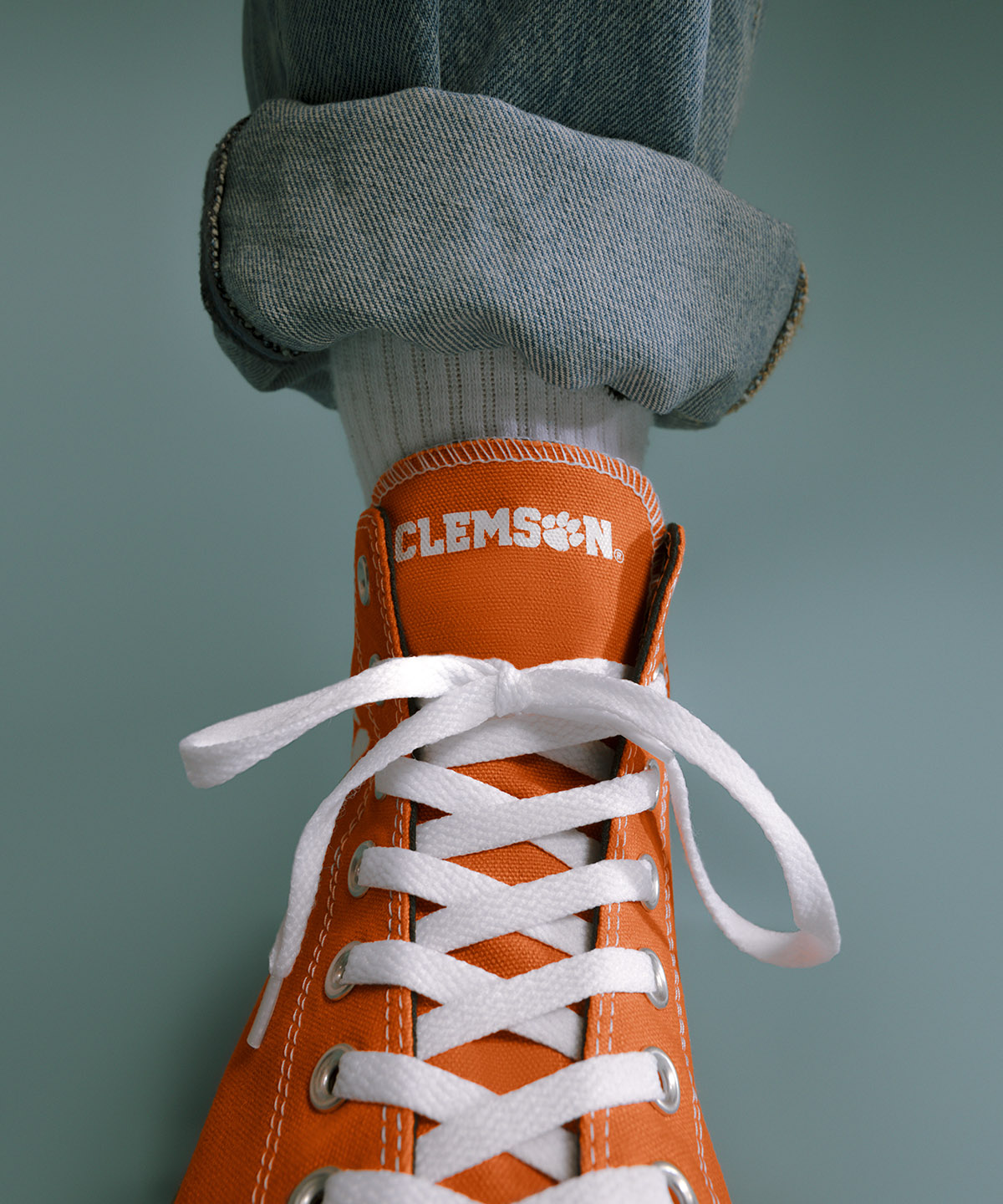 a up close view of a converse shoe with the word clemson under a cuffed blue jean with a blue background