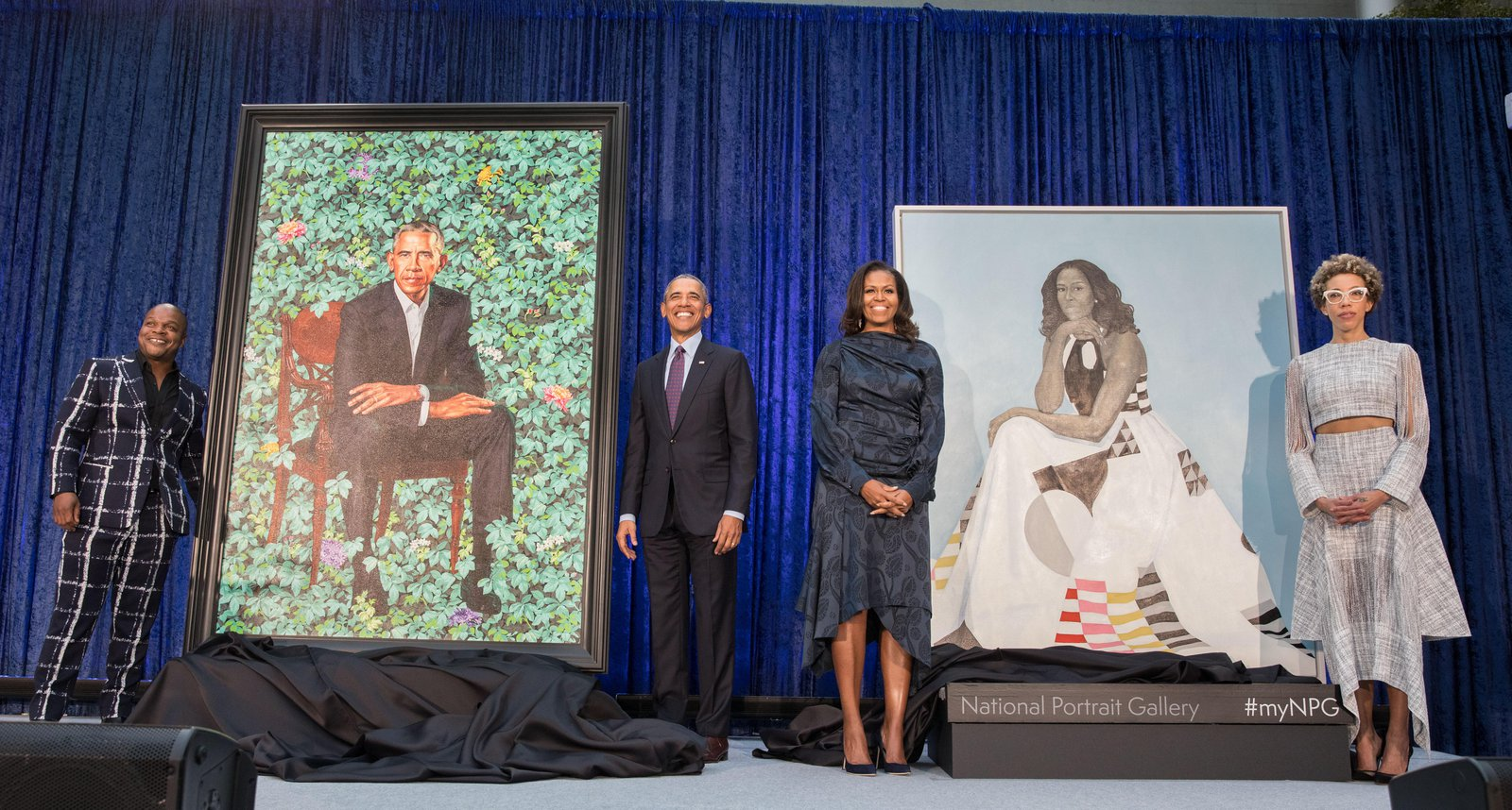 Barack and Michelle Obama stand center stage, smiling. Beside each is their respective portrait. Beside each portrait is the portrait's painter. Background is a dark blue curtain.