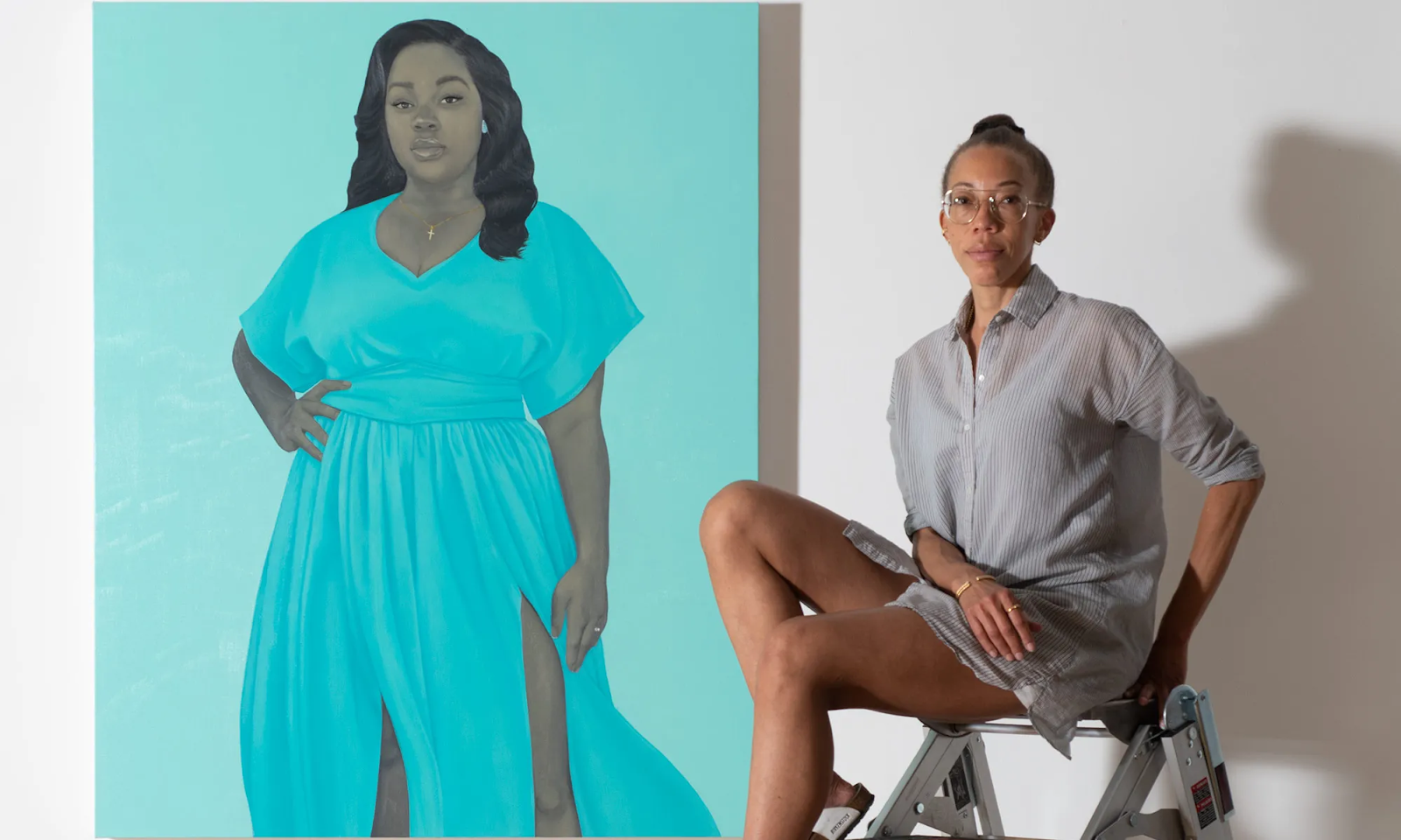 Amy Sherald sits on the top of a ladder in a button down shirt next to her portrait of Breonna Taylor. Breonna wears a bright blue dress and stands out in black and white against a lighter blue canvas. She has her right hand on her hip and wears a gold cross necklace. Sherald's pose mirrors Breonna's; her left elbow is bent at her side to support her on the ladder. Both women look at the camera with a barely-there smile.