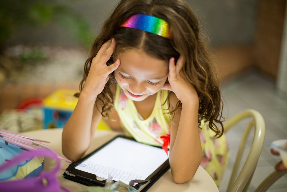 Child wearing a rainbow headband smiles down at a tablet on the table in front of her.