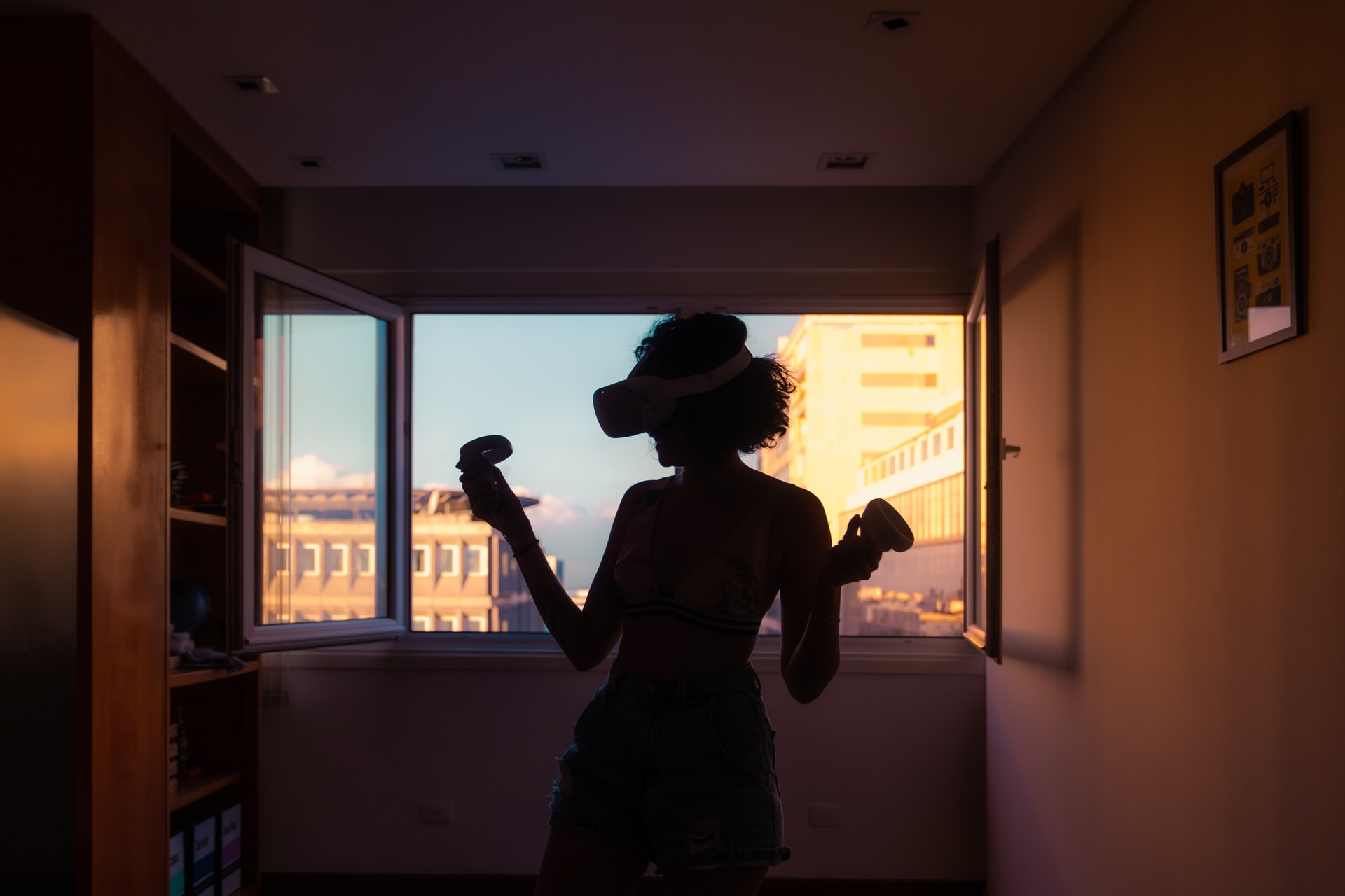 Woman wearing a VR headset is silhouetted in front of a window pouring golden light onto a cityscape.