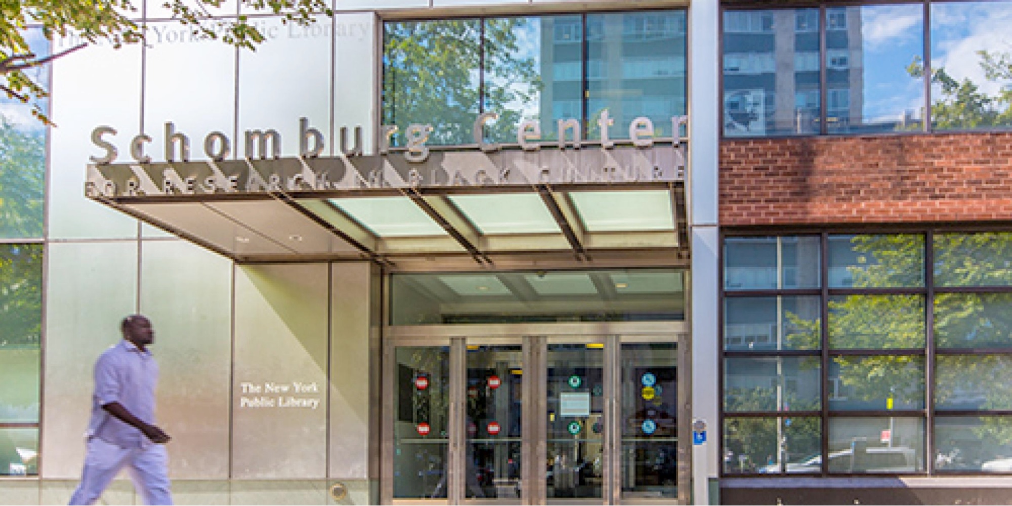 Image of a window-covered building taken from across the street.. Sign above the glass doors reads "Schomburg Center for Research in Black Culture." On the bottom left of frame, a man in lavender walks beside the building. It's a sunny day. A tree is reflected in the building's many windows.