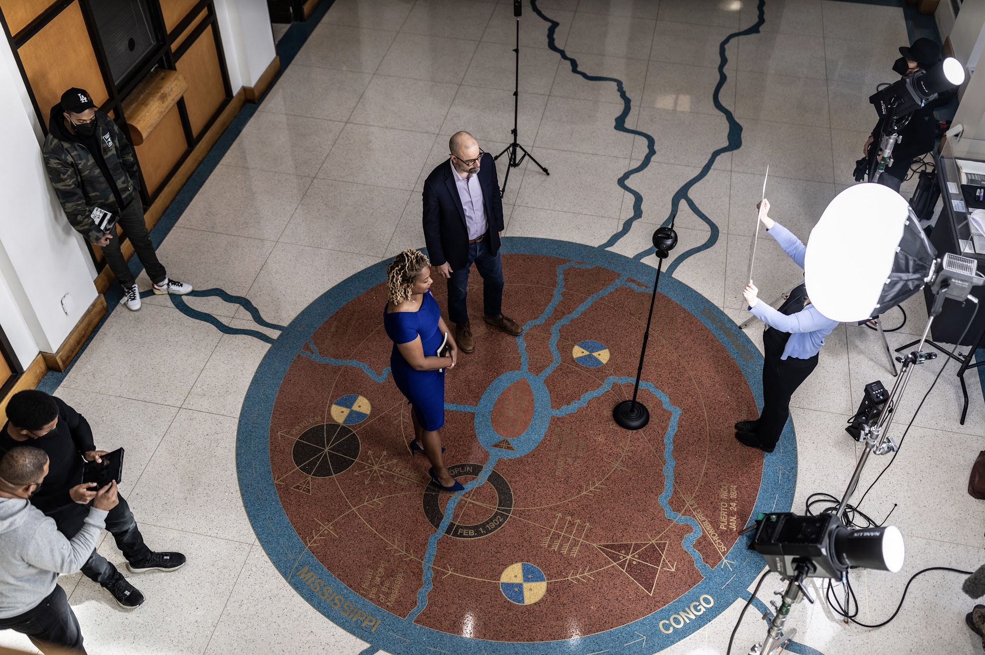 Birds eye view of set. Kai and Schomburg Center representative stand on circular emblem (the cosmogram) on the ground. The cosmogram is bronze-colored. Blue rivers run through it and then off across the white tiled floor. Crew members and equipment are visible around the emblem.