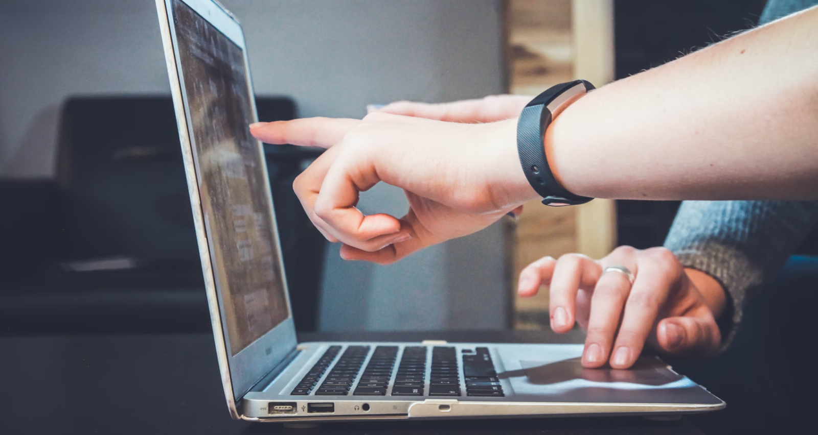 Side view of open laptop. Hand wearing a fit watch points to it. Hand of another person scrolls on the trackpad.