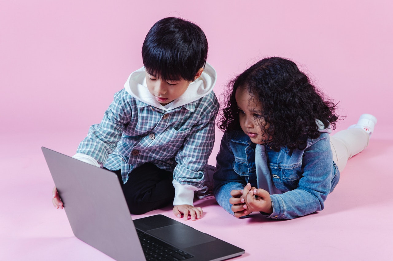 Two kids watching computer in studio. Light lavender pink background.