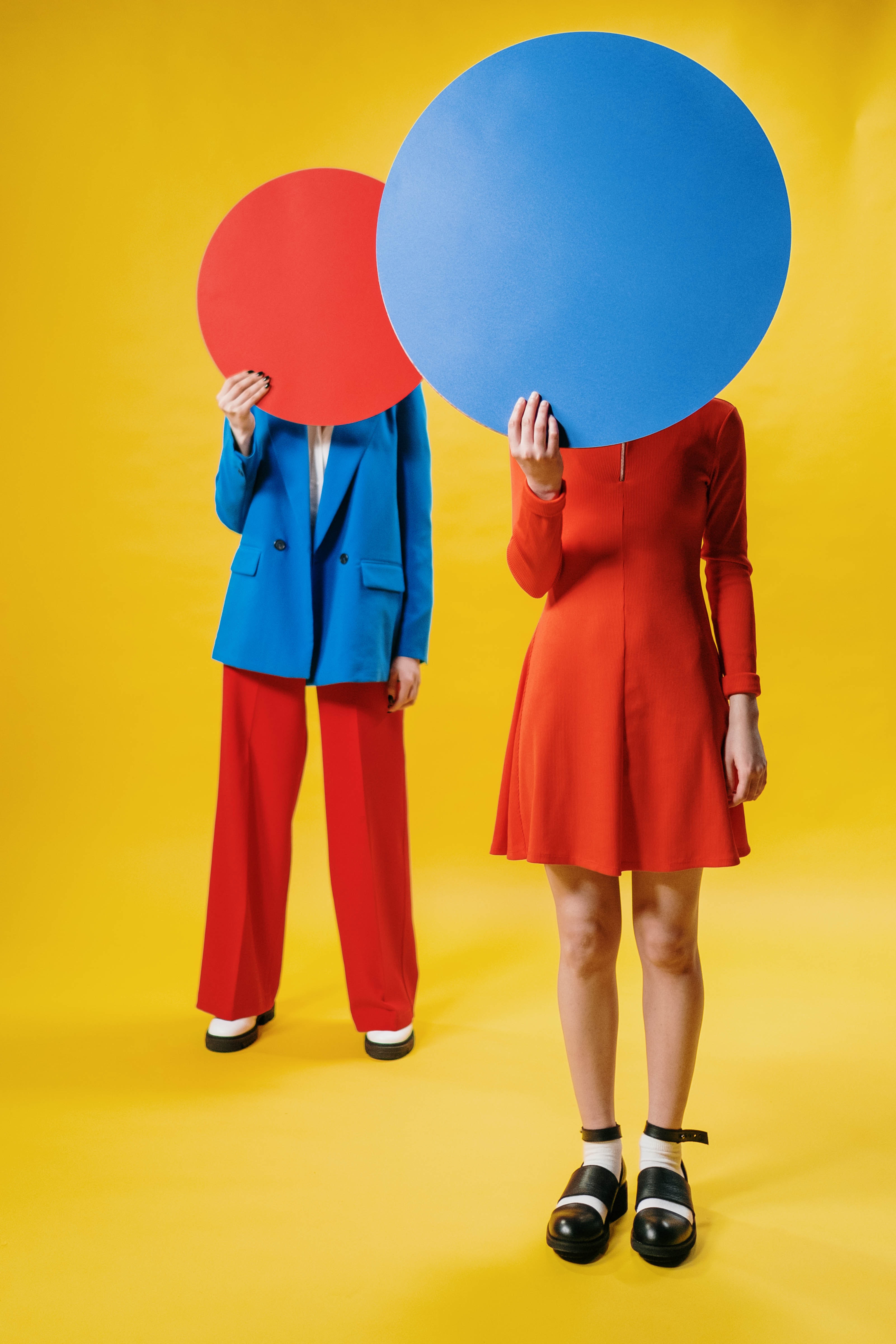 we have two people dressed in bright reds and blues on a bright yellow background, they are holding big circles in front of their faces the person dressed in a red dress is holding a blue circle, the person dressed in red slacks and a blue blazer is holding a red circle