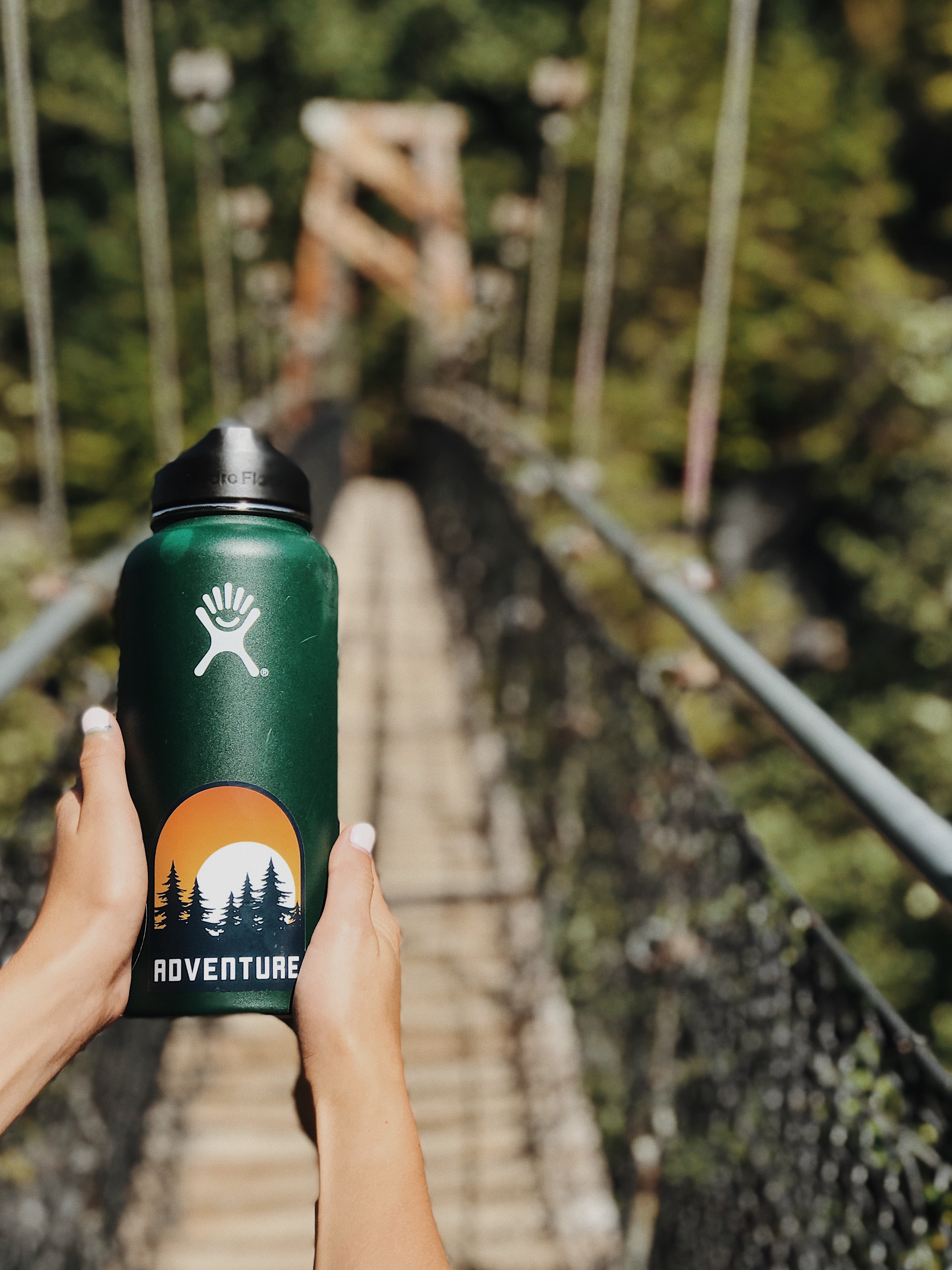 we are on a bridge in the middle of the forest, all we can see is the hands of a person holding a hydroflask waterbottle in a forest green color with a sticker that has a sunset behind the trees that says adventure
