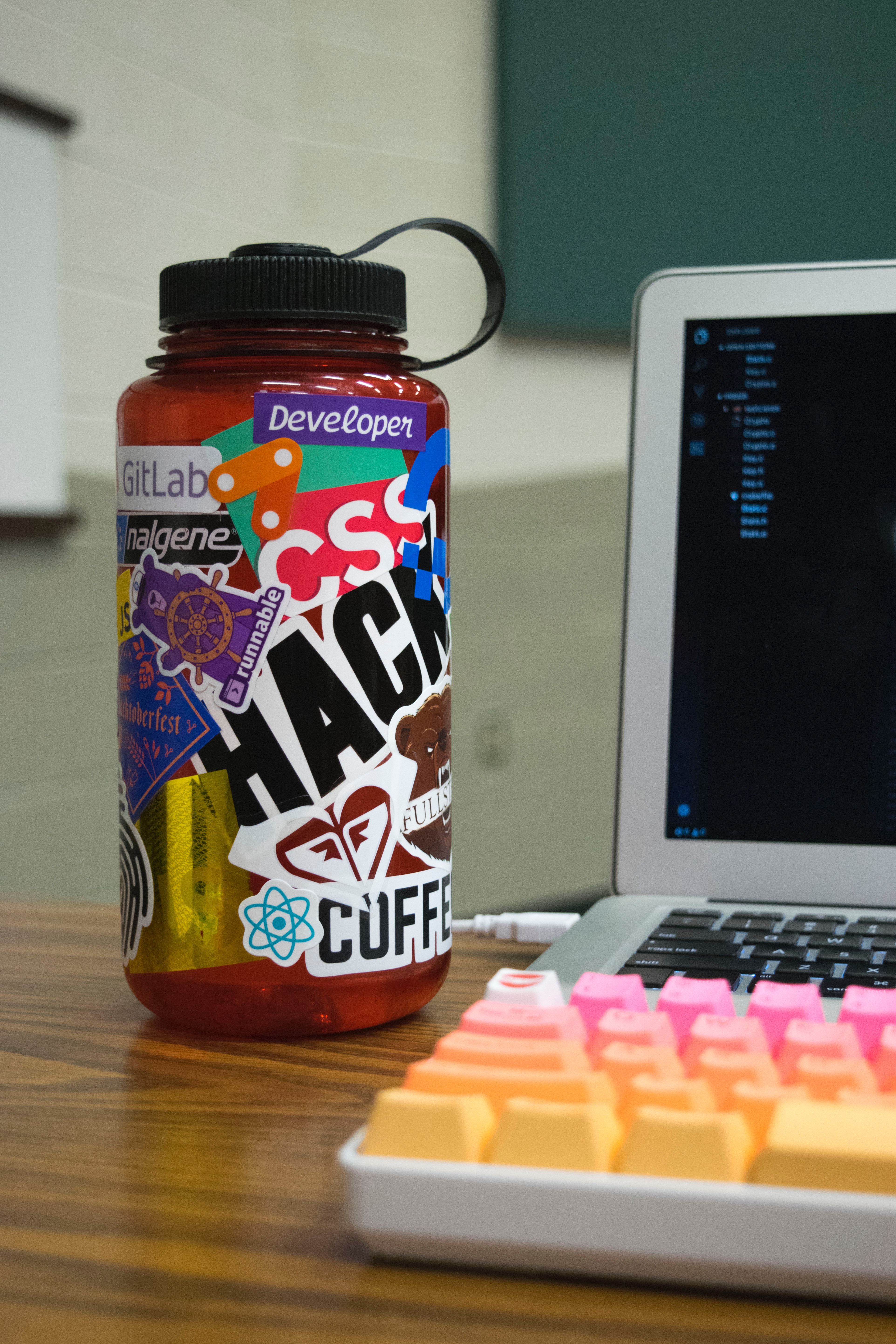 there is a nalgene water bottle covered in stickers sitting on someones desk, they have a laptop open to the side of the bottle, but it is out of focus 