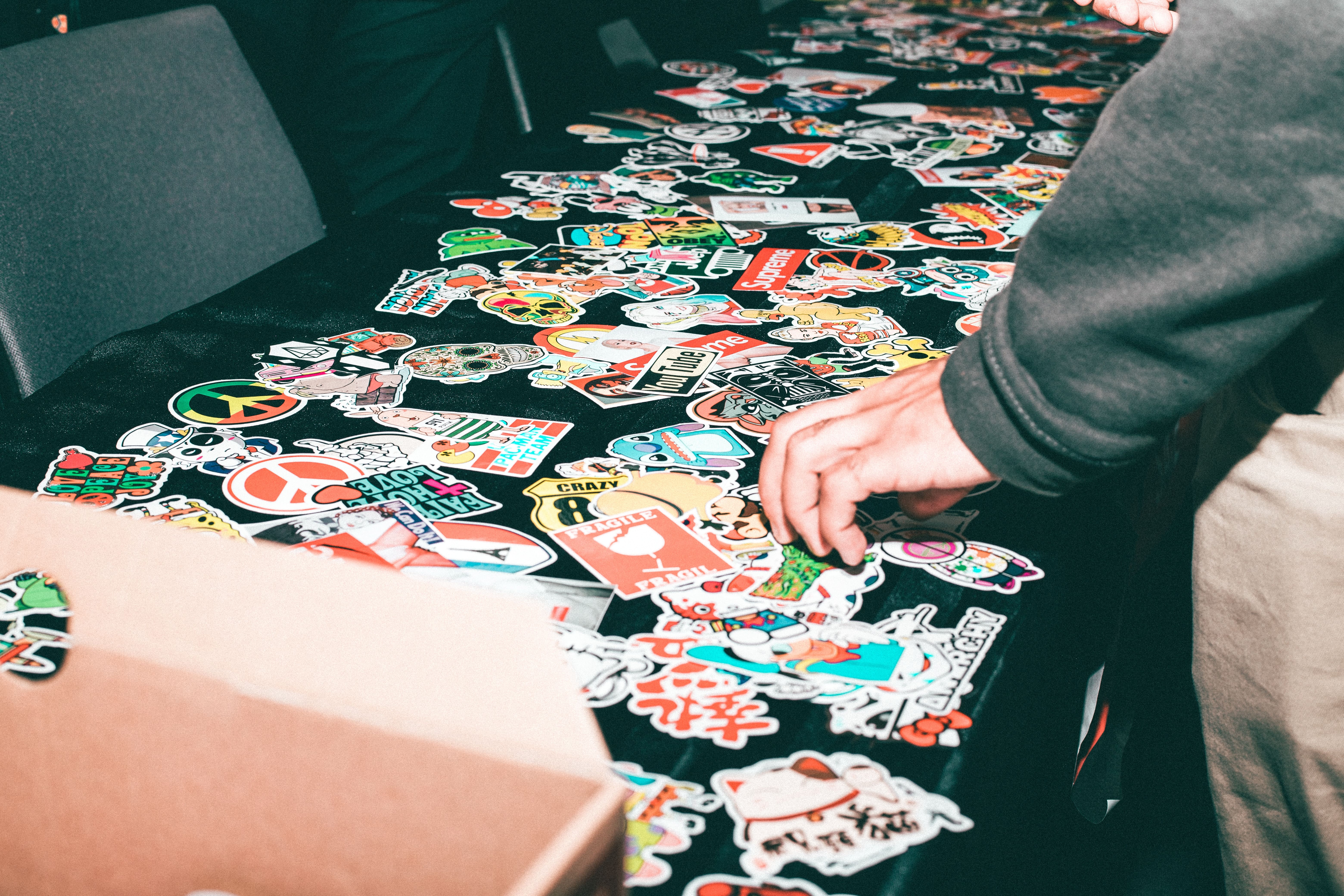 a table with a lot of stickers just covering it and scattered about, and a hand reaching to grab a sticker