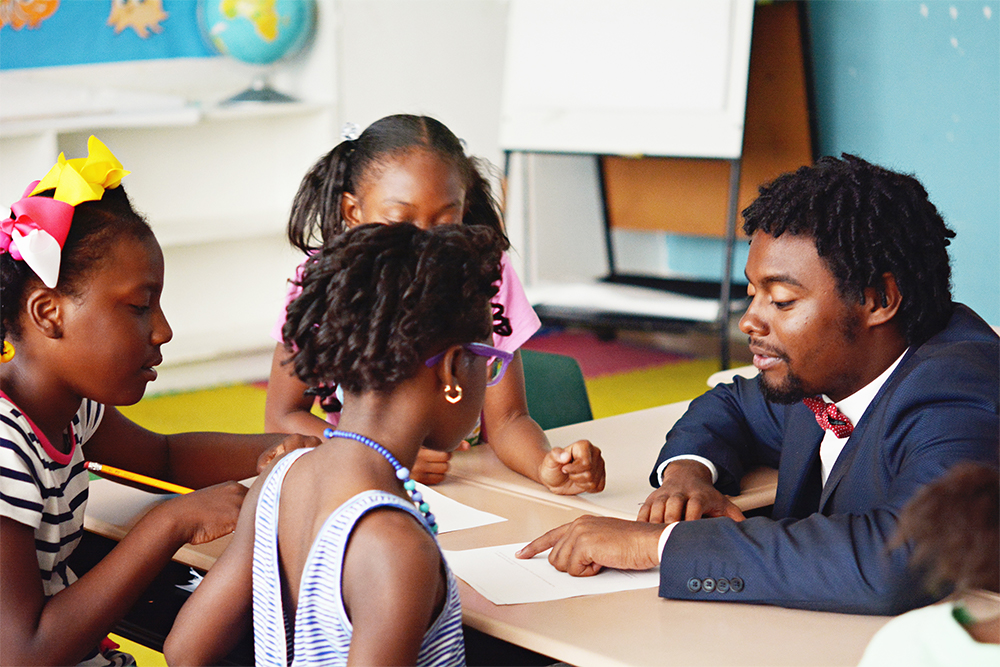 Teacher seated at desk with students, helping with their work.
