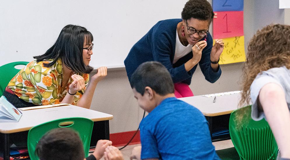 Two teachers in front of class, smiling as they teach.