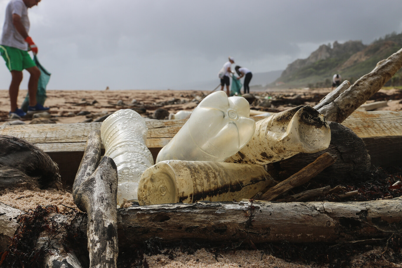 Plastic waste in the foreground, a beach in the background, a person walking past.