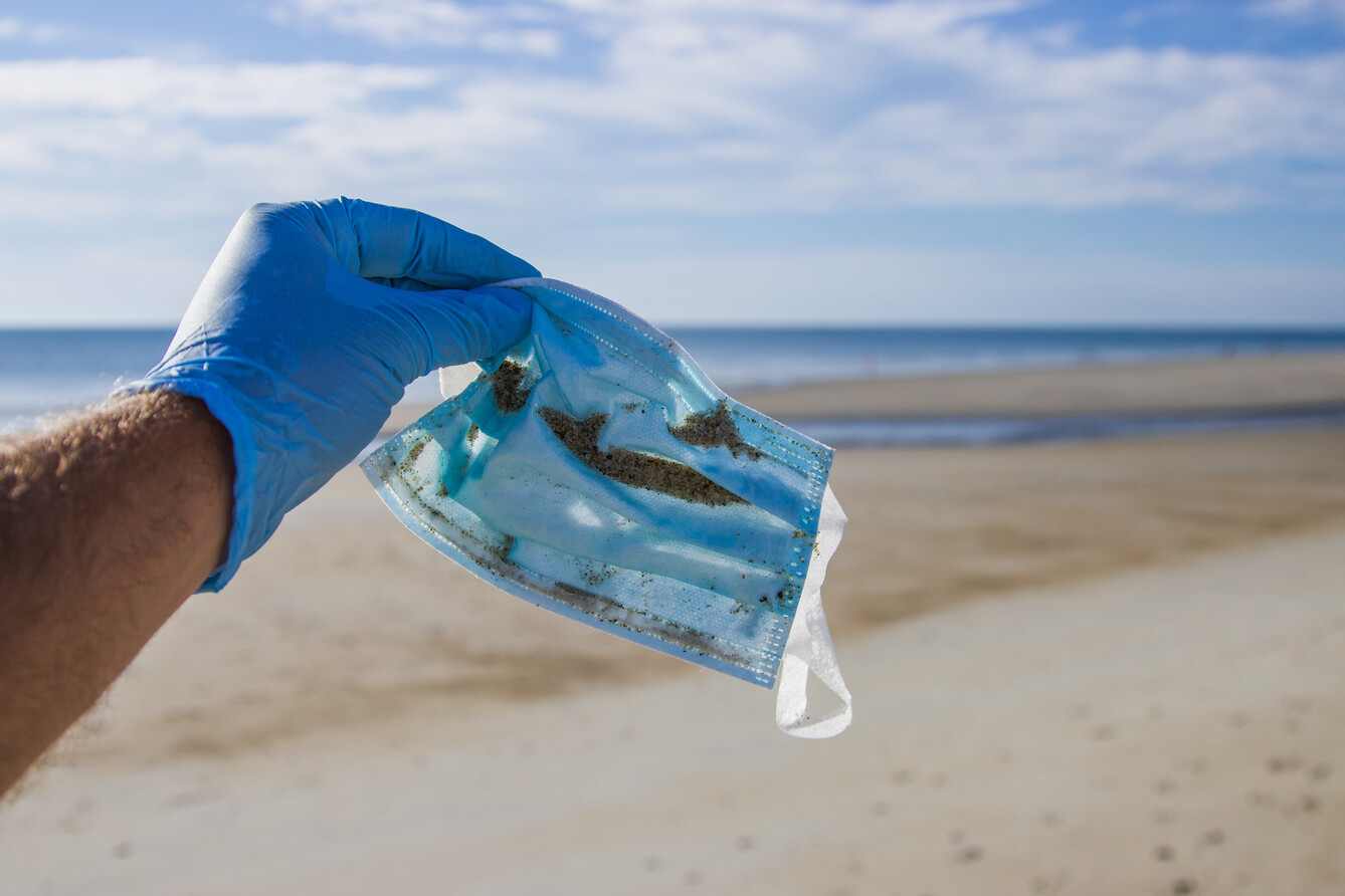 A plastic-gloved hand holding a medical face mask, with a beach in the background.
