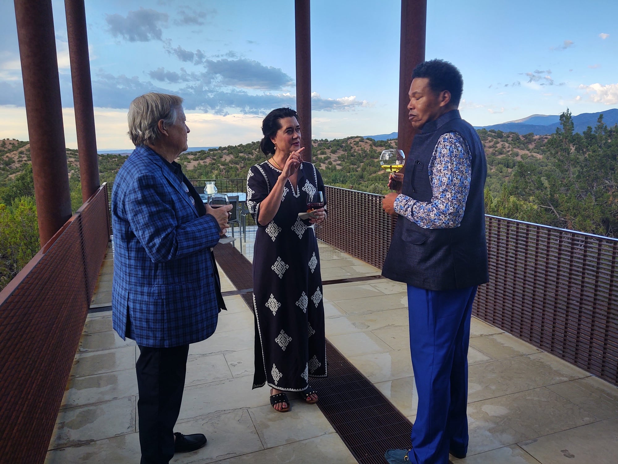 Group of three diverse donors, conversing, holding wine on a terrace overlooking the Tesuque landscape