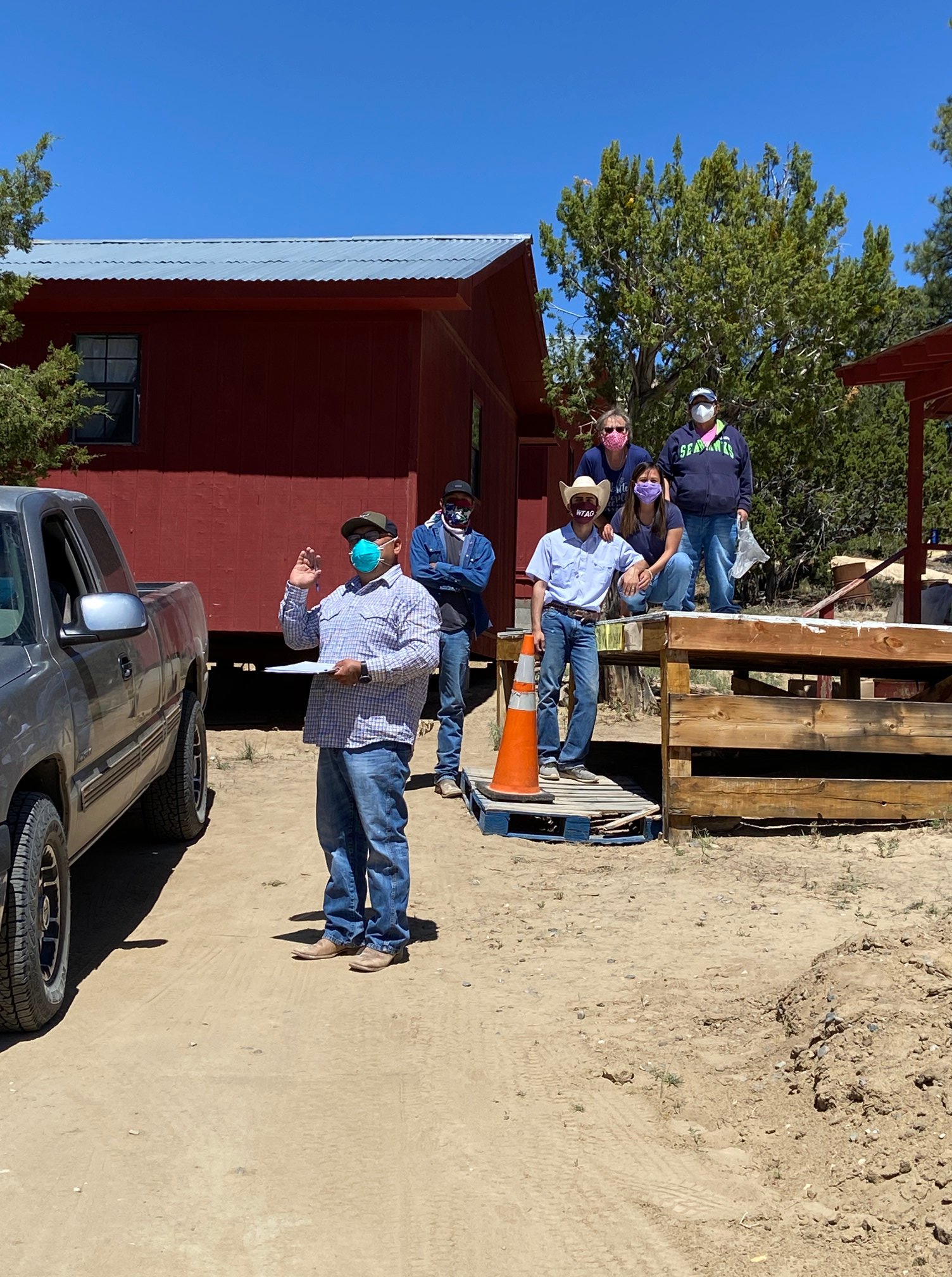 Group of diverse volunteers next to a truck and in front of a red buidling wearing masks, man in front is waving carrying clipboard is waving