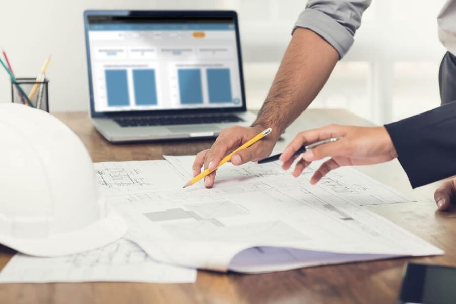 Deux mains de personnes qui dessinent sur un plan de chantier avec un ordinateur en fond
