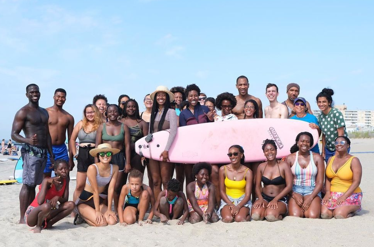 Terumi Murao surrounded by group of people holding a pink surfboard at the beach