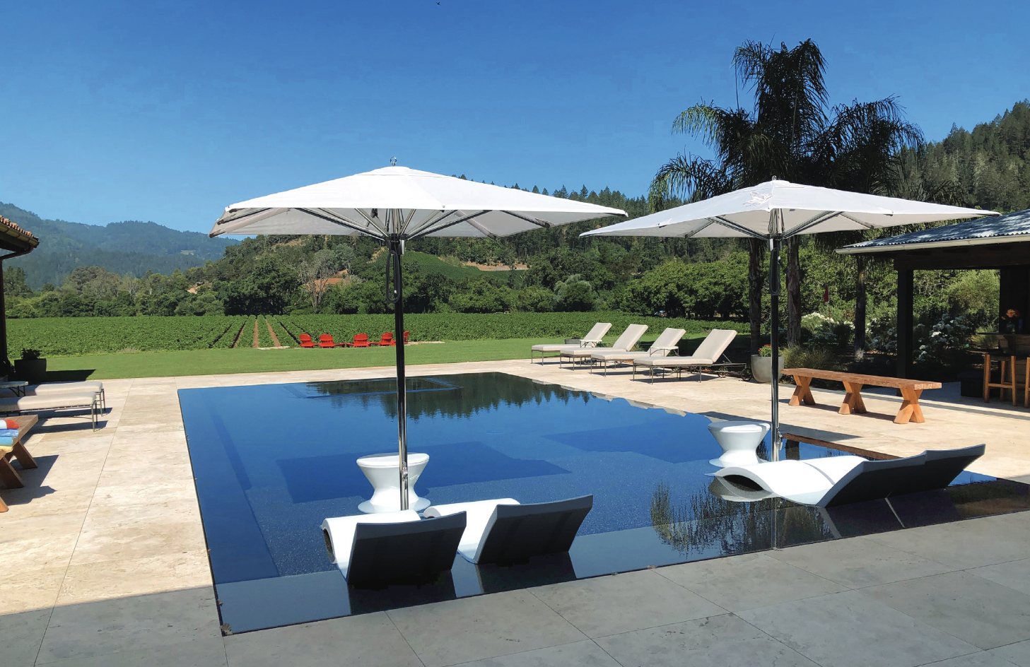 A pool sits sunken in a cream stone patio, with modern loungers and a view of lush greenery