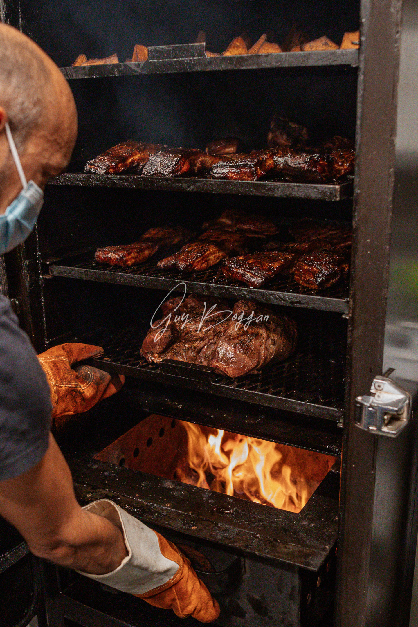 A man with an oven glove on opening the bottom fire engulfed drawer of a meat smoker, meat is cooking on racks above 