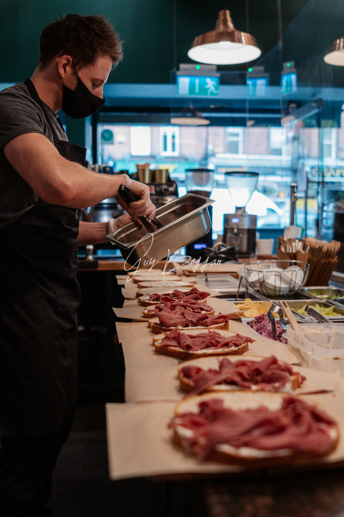 A man lays meat onto multiple open half made sandwiches behind a deli counter