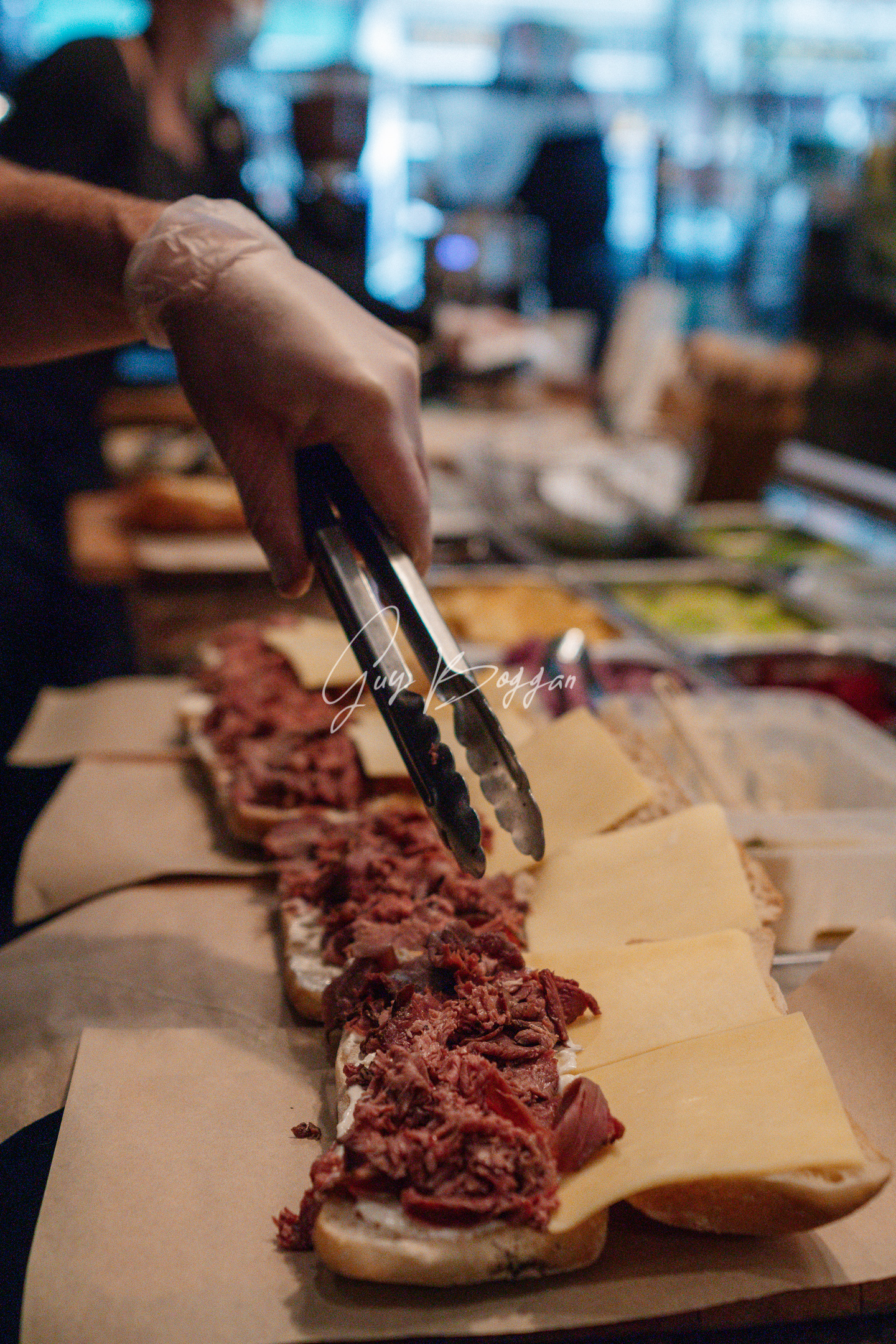 A close up of meat being placed onto multiple open half made sandwiches behind a deli counter