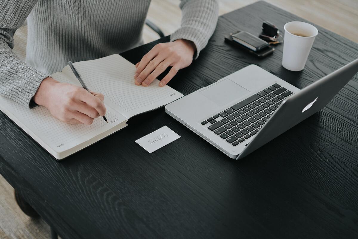 person-writing-on-white-notebook-with-laptop-on-table