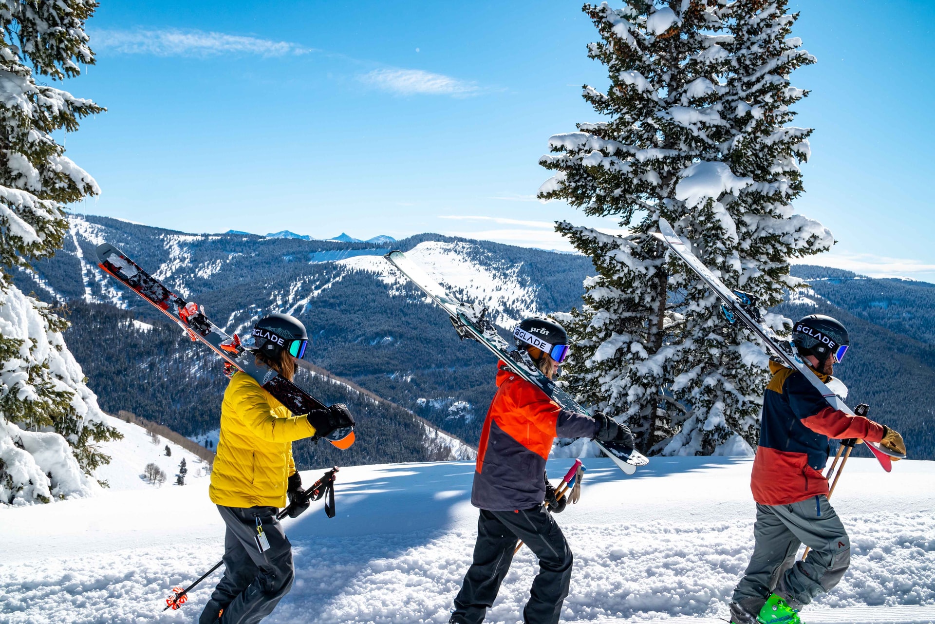 Three people dressed in ski gear walking through the snow carrying their ski's on their shoulders.