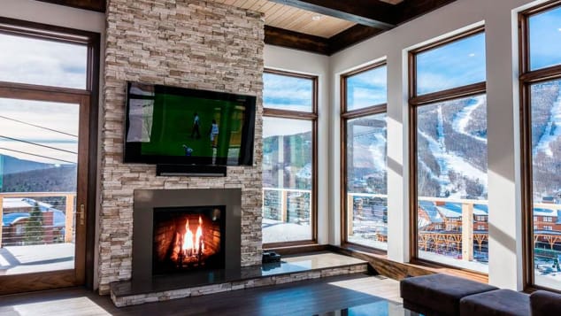 A luxurious modern ski lodge with wall-to-wall windows, a stone fireplace and TV mounted above the fireplace.
