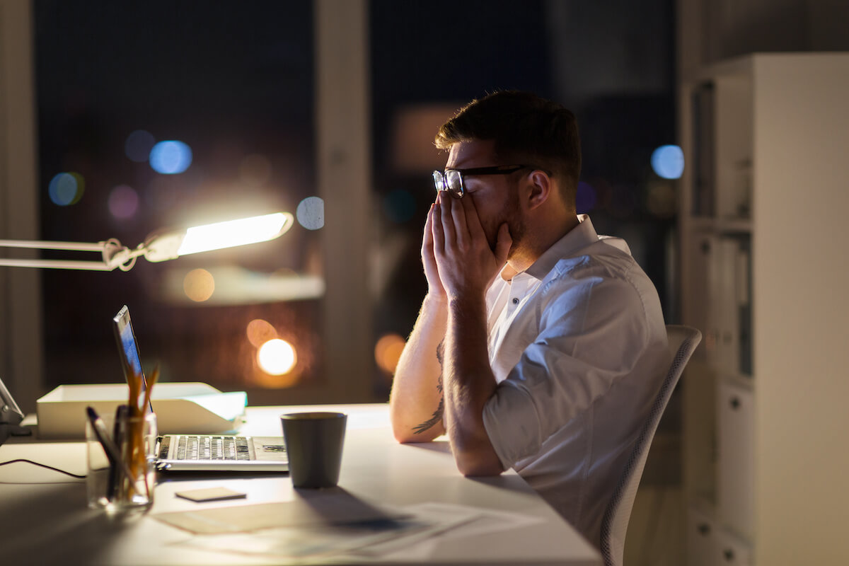 Context switching: A tired, stressed man rubs his eyes while sitting at a desk with his laptop late at night
