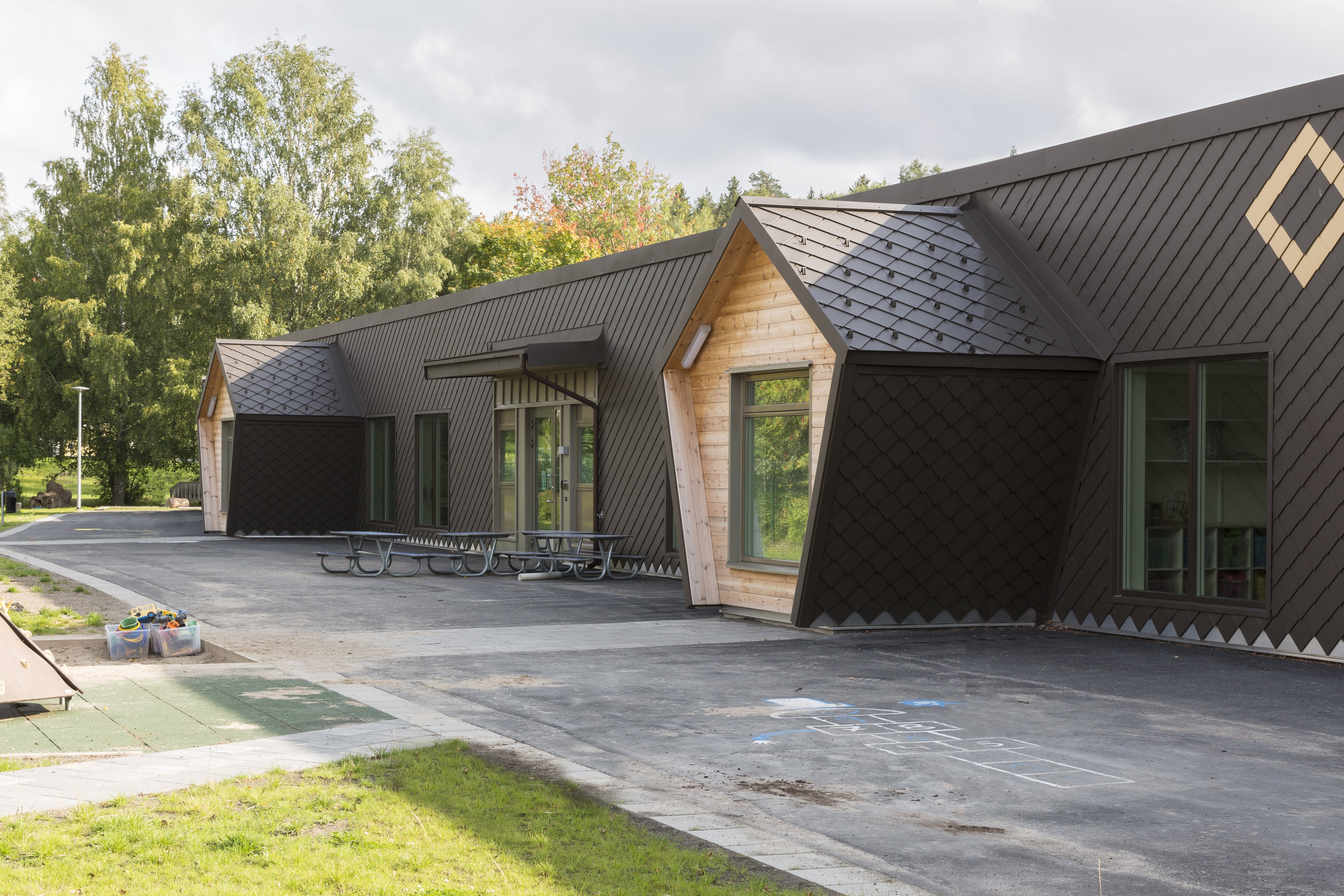 Entré med hexagonformade burspråk mot rastgård med lekplats och sandlåda.