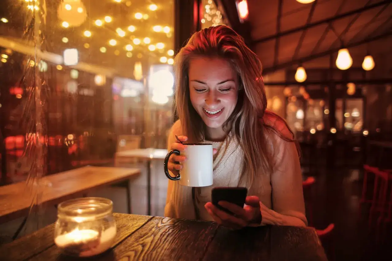 Close-up of a woman smiling while holding a steaming cup of coffee, looking at her mobile phone.