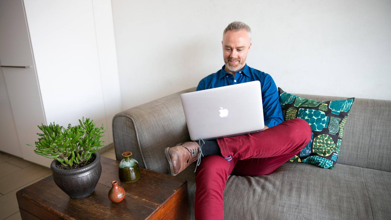 A man sitting on a couch, with a laptop on his lap. He has one leg crossed over the other.