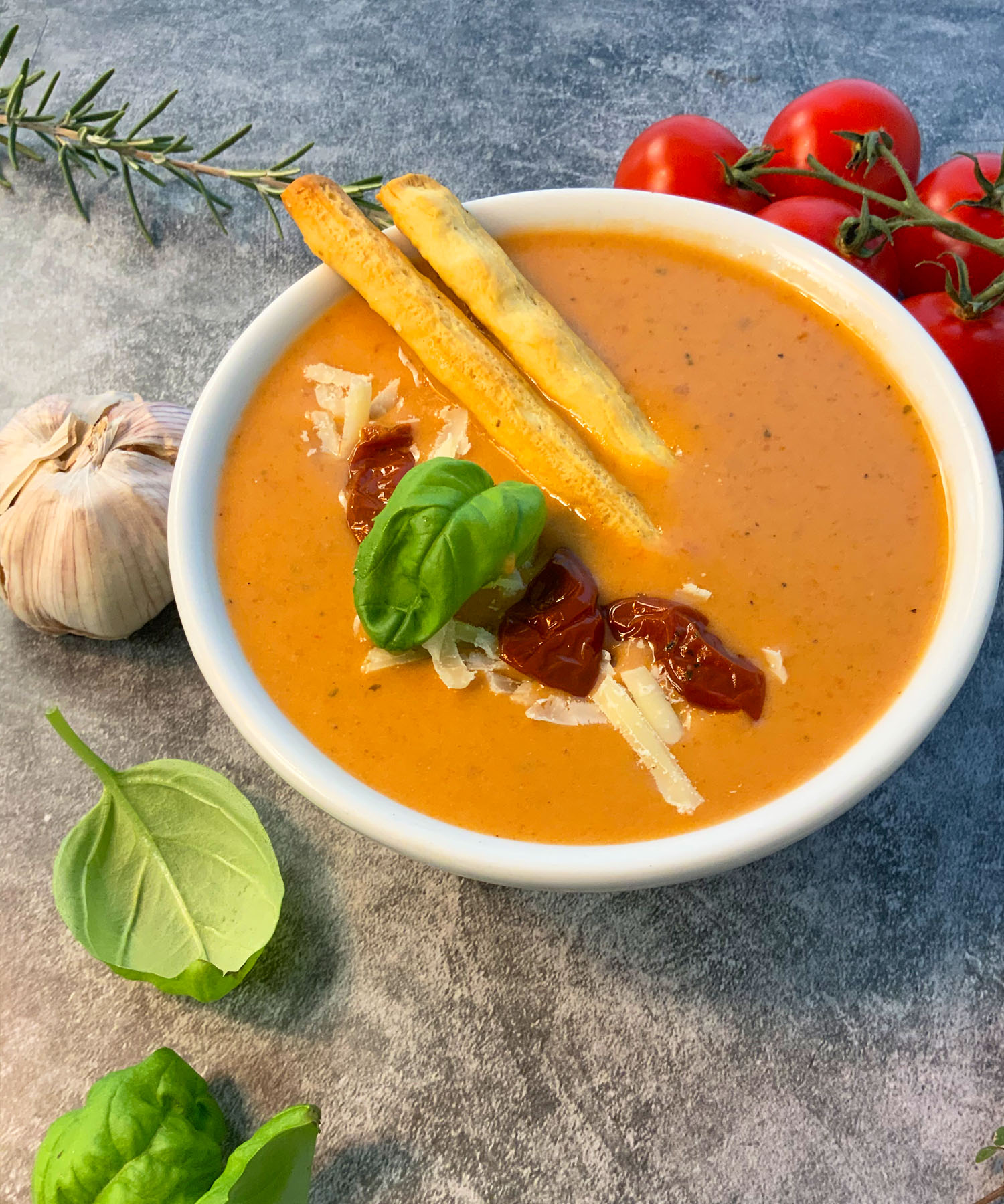 Looking down at a bowl of warm tomato soup with fresh vegetables laying around the bowl.
