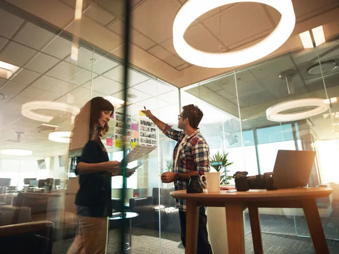 Two people in an office with glass walls adding post it notes to the wall