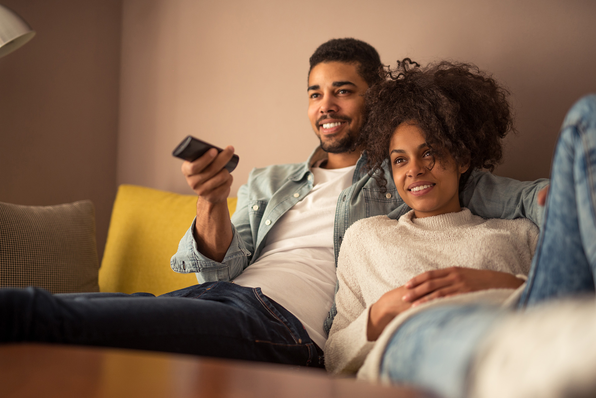 A male and female sat on a sofa watching tv.