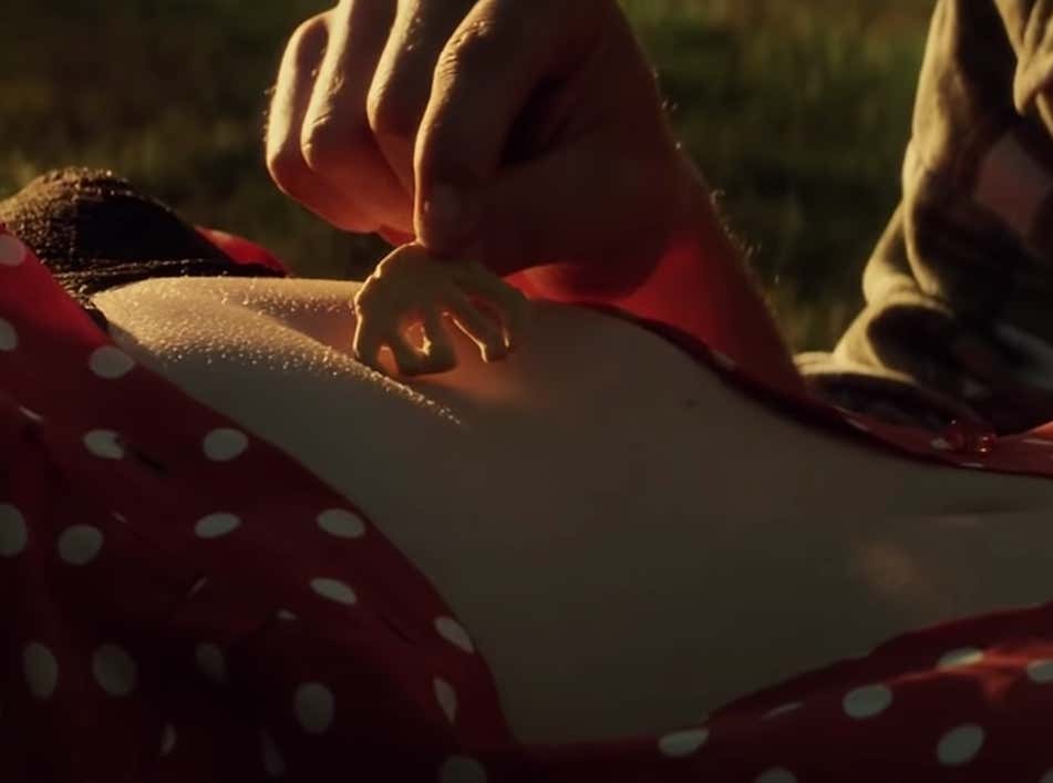 Film still from Armageddon. A man's hand walks an animal cracker against a woman's bare torso, down to her belly button. The sun shines down on her skin.