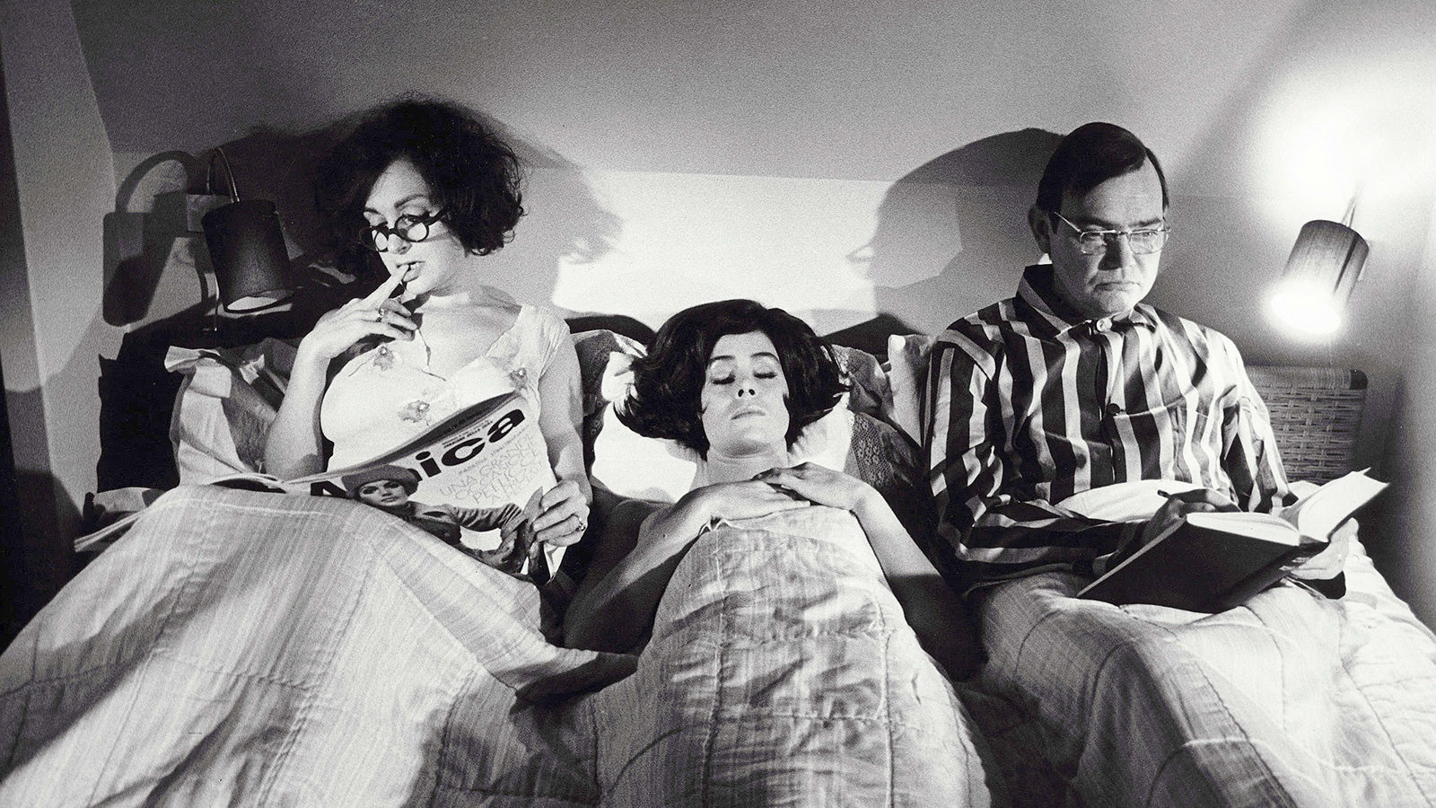 Film still from Duet For Cannibals. Two women and a men share a bed. Two of them are reading, while the woman in the middle lies asleep.